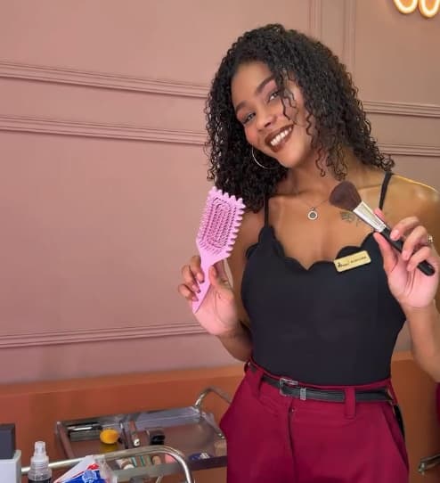 Ana Clara, de cachos escuros e definidos, sorrindo com uma escova rosa numa mão e um pincel de maquiagem na outra.
