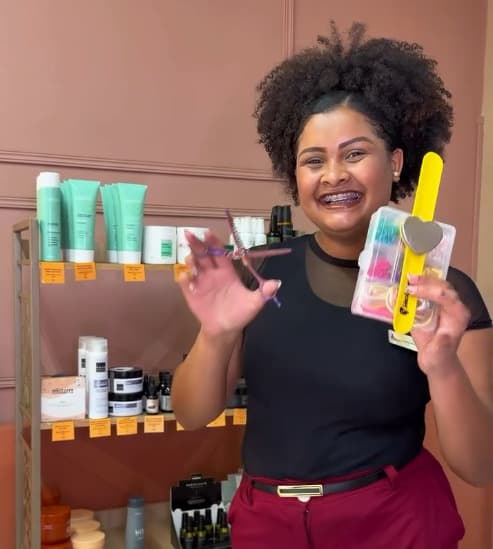 Mikaellen Granda, de cabelo crespo preso no alto, sorrindo com uma tesoura na mão.