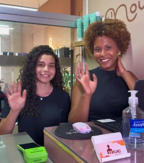 Carine Dolatese, à esquerda, e Estelma Ribeiro acenando atrás do balcão da recepção da Miaxa Cachos.