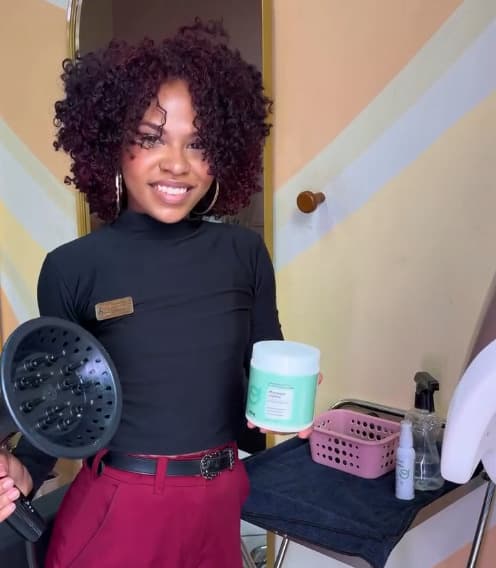 Roberta Vitória, de cachos volumosos em tom vinho, segurando um difusor e um pote de creme.