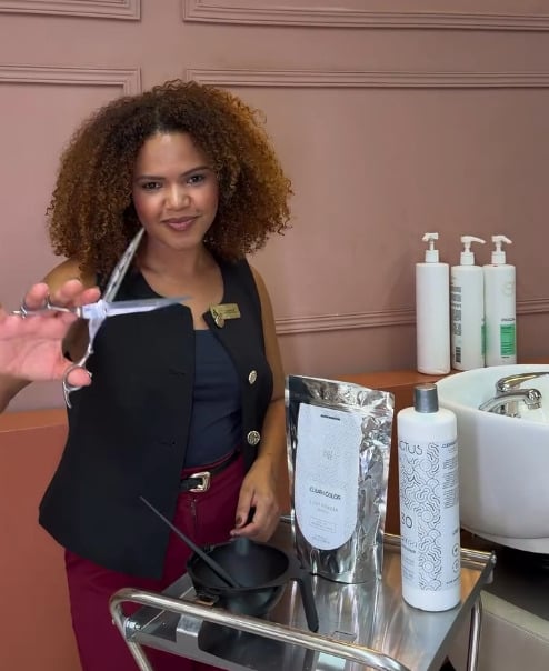 Mailane Lourenço, fundadora da Miaxa, de cachos volumosos com mechas cor de mel, segurando uma tesoura aberta no salão.